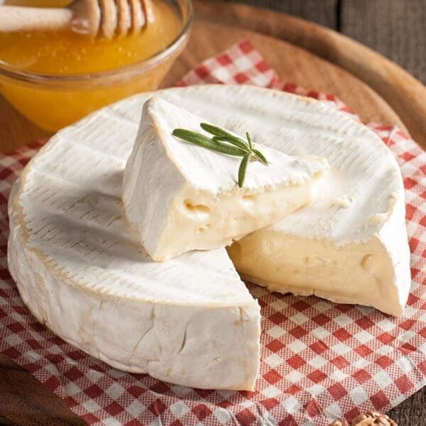 Soft cheese wheel with white rind, sliced to show creamy inside, on checkered cloth with honey.
