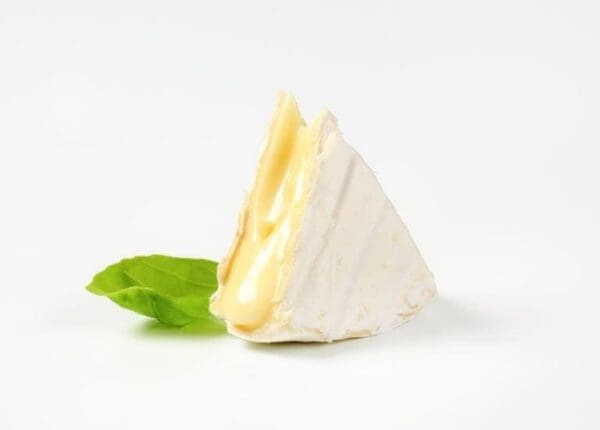 Wedge of soft cheese with white rind on a surface, with a green basil leaf beside it.
