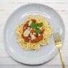Plate of spaghetti with meat sauce, parsley, parmesan; gold fork on white wooden surface.