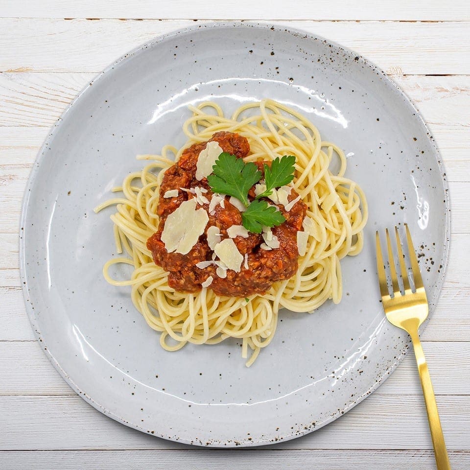 Plate of spaghetti with meat sauce, parsley, parmesan; gold fork on white wooden surface.