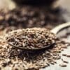 Spoonful of cumin seeds with scattered seeds on blurred background surface.