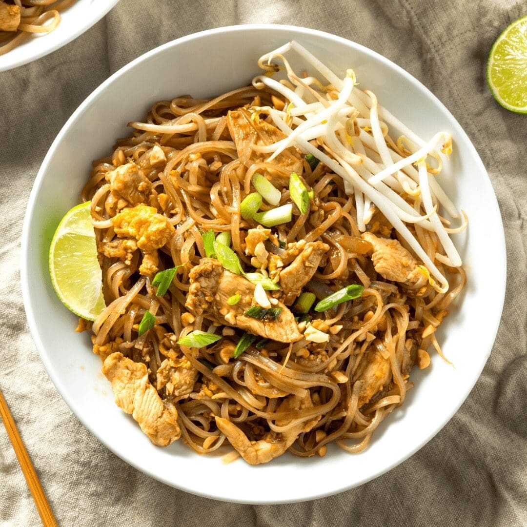 Bowl of stir-fried noodles with chicken, bean sprouts, green onions, peanuts, and lime wedge.