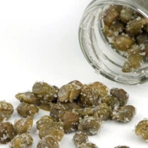 Glass jar tipped over with salted capers on a white surface.