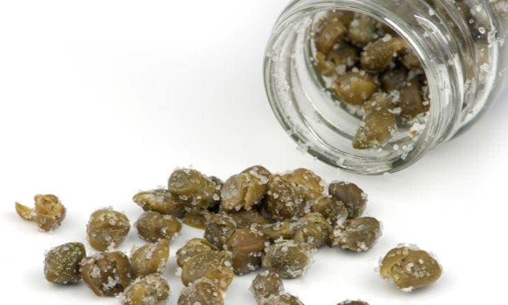 Glass jar tipped over with salted capers on a white surface.