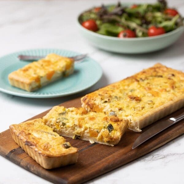 Rectangular vegetable quiche sliced on board, piece on turquoise plate, salad in background.