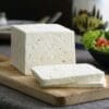 White cheese block on cutting board, with salad, olives, and container in the background.