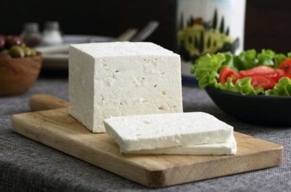 White cheese block on cutting board, with salad, olives, and container in the background.