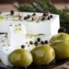 Plate with white cheese, herbs, peppercorns, green olives, and sprig of herbs in background.