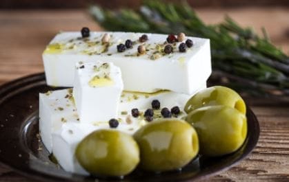 Plate with white cheese, herbs, peppercorns, green olives, and sprig of herbs in background.