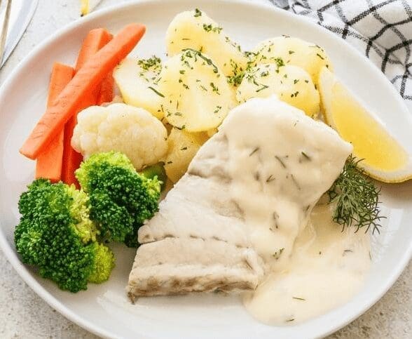 Plate with white fish in creamy sauce, boiled potatoes, lemon, broccoli, carrots, and cauliflower.