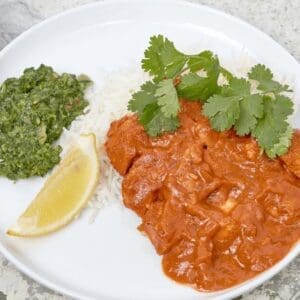 Plate of white rice with red curry, cilantro, green chutney, and a lemon wedge.
