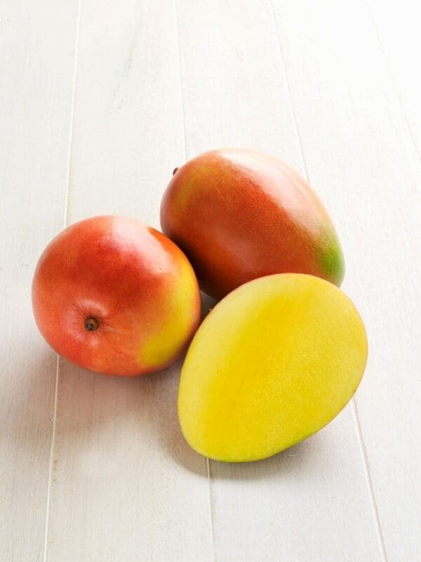 Two whole mangoes and one sliced, revealing bright yellow flesh on light wooden surface.