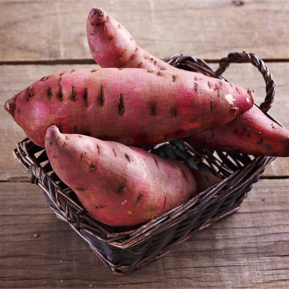 Wicker basket with large reddish-brown sweet potatoes on wooden surface.