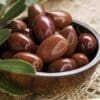 Wooden bowl of glossy brown olives on tan fabric, surrounded by green leaves.