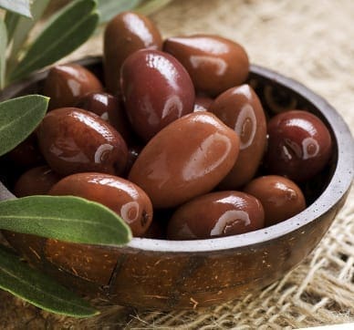 Wooden bowl of glossy brown olives on tan fabric, surrounded by green leaves.