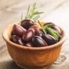 Wooden bowl with dark olives and rosemary on a wooden surface.