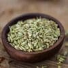 Wooden bowl of dried, greenish fennel seeds on rustic wooden surface, some scattered around.