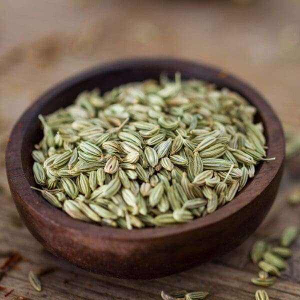 Wooden bowl of dried, greenish fennel seeds on rustic wooden surface, some scattered around.