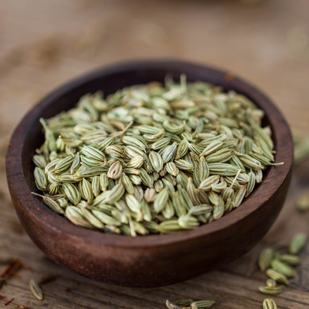 Wooden bowl of dried, greenish fennel seeds on rustic wooden surface, some scattered around.