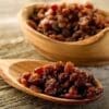 Wooden spoon and bowl with dried raisins on a wooden surface.