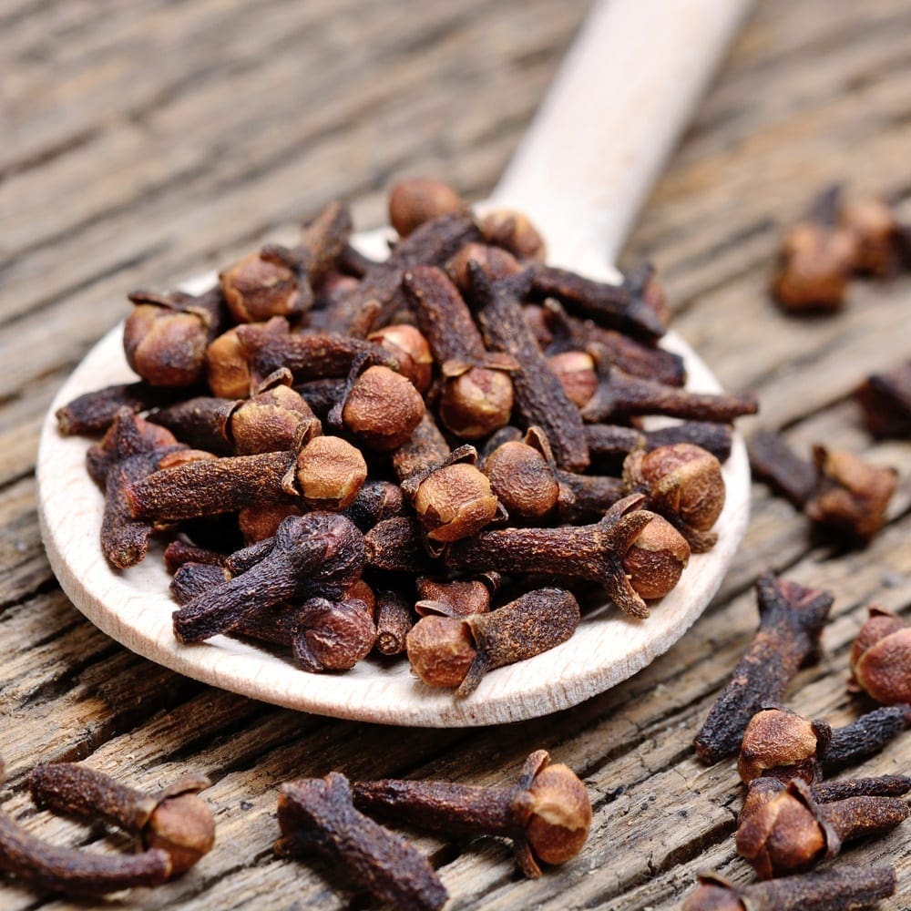 Wooden spoon filled with dried cloves on rustic wooden surface, scattered cloves nearby.